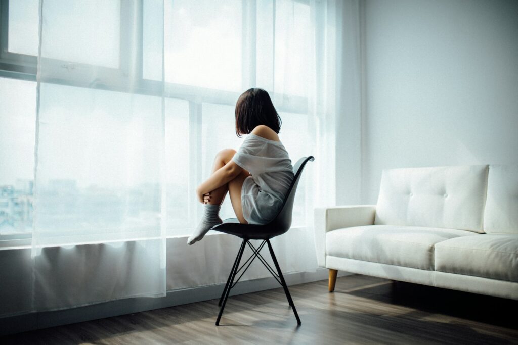 femme assise sur une chaise noire devant une fenêtre en verre avec des rideaux blancs et souffrant de dépression saisonnière
